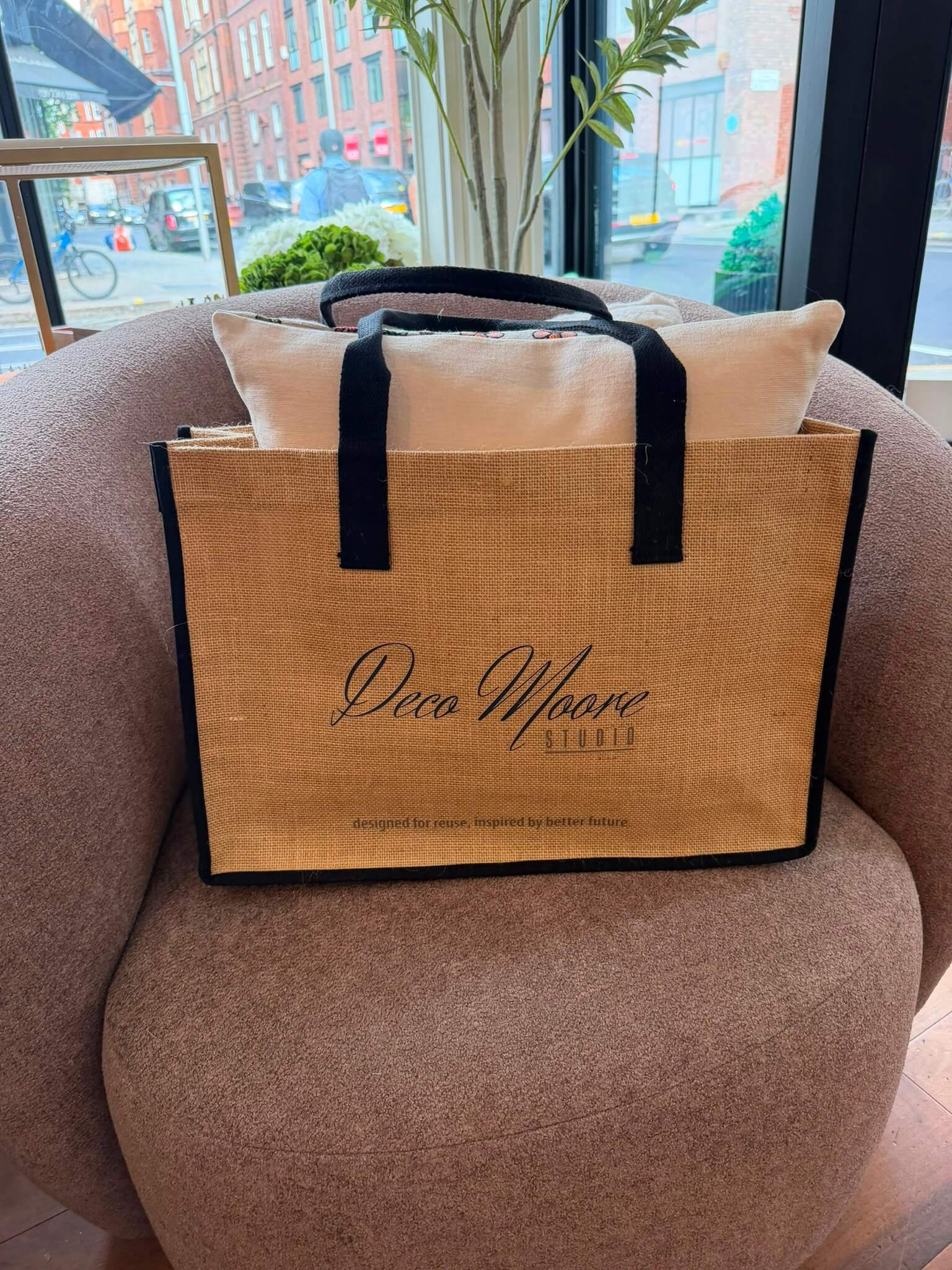 Deco Moore Jute Tote Bag displayed on a chair with a pillow, featuring strong handles and logo for stylish shopping.