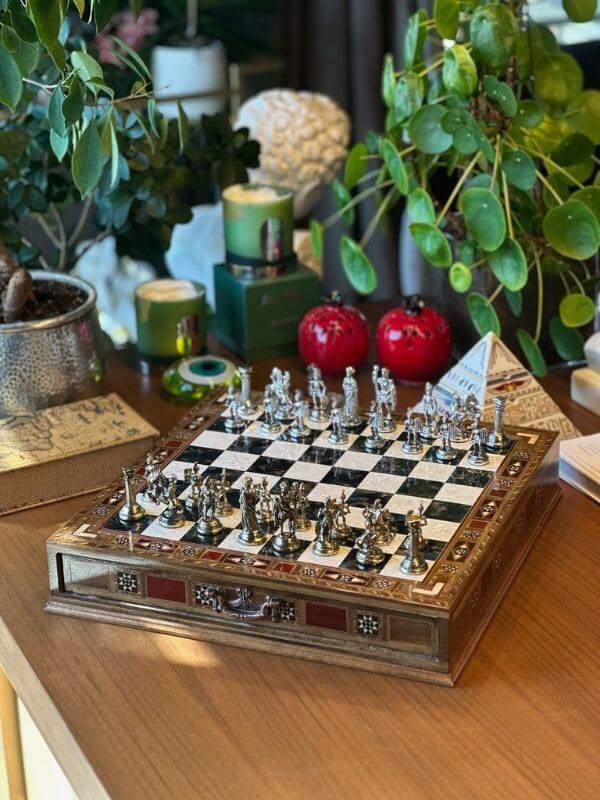 Large handmade chess set displayed on a wooden table surrounded by plants and decorative items.