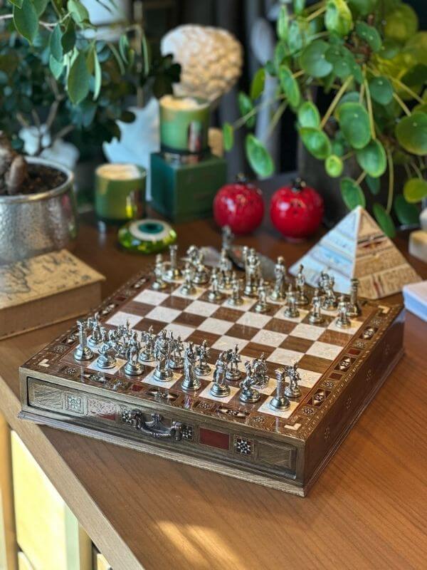 Large handmade chess set displayed on a wooden table surrounded by plants and decorative items.